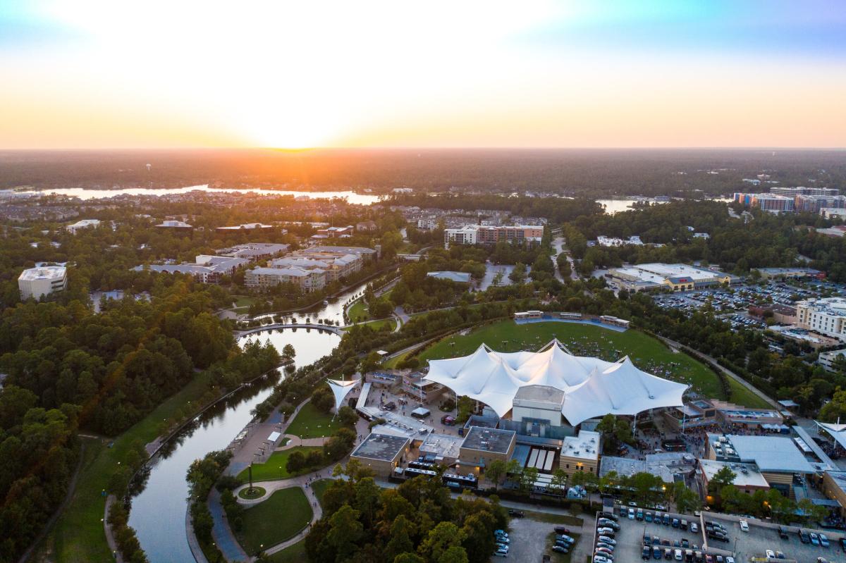 An aerial view of The Woodlands, Texas, featuring the Pavilion in the lower right of the frame. The Waterway wraps around the Pavilion from the left, leading into various business and residential areas before reaching Lake Woodlands. The sun is setting beautifully on the horizon.