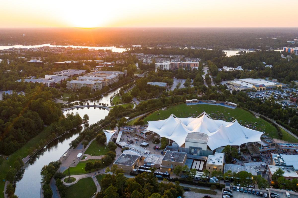 An aerial view of The Woodlands, Texas with a focus on The Cynthia Woods Mitchell Pavilion. The Waterway wraps around the Pavilion from the left into business and residential areas. The sun is setting beautifully on the horizon.