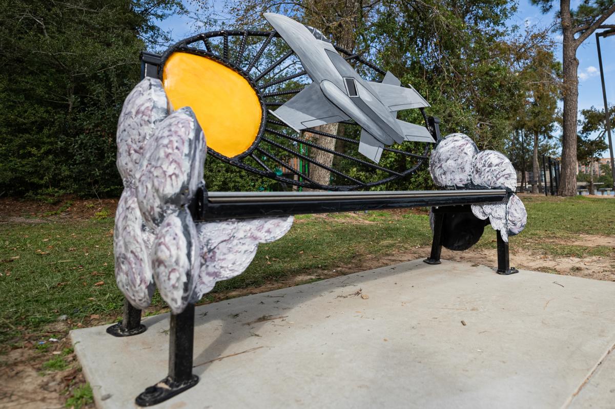 The back of the bench includes a yellow circle with thin, black, metal arms branching outward the center, representing the sun and its rays. An image of a large gray jet soars over the sun. The arms of the bench are shaped like clouds, painted in various shades of gray with maroon accents.