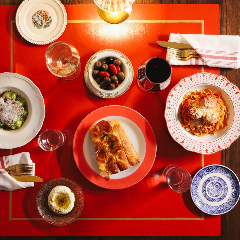 A red table is set with two place settings. One has a pasta with red sauce, the other a pasta with pesto and cheese. A bowl of olives and a plate of bread are between the two settings.