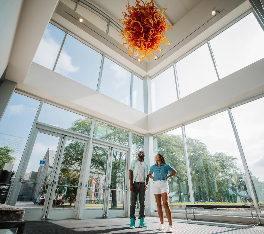 A man and a woman stand underneath a glass-blown sculpture located in the entry of the Kalamazoo Institute of Arts.