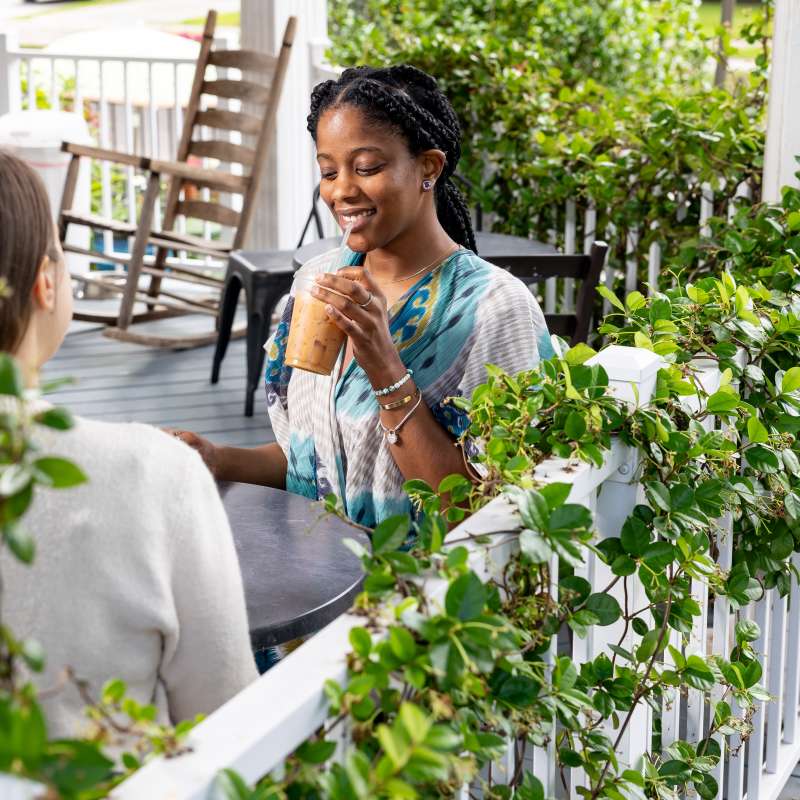 Two women enjoy iced drinks on a vine-wrapped front porch at Curate Essentials in Fayetteville, NC, surrounded by greenery and rocking chairs.