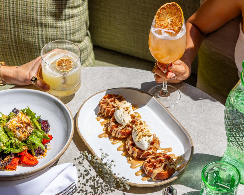 A plate of carrot cake waffles sitting on a table surrounded by two hands holding cocktails.