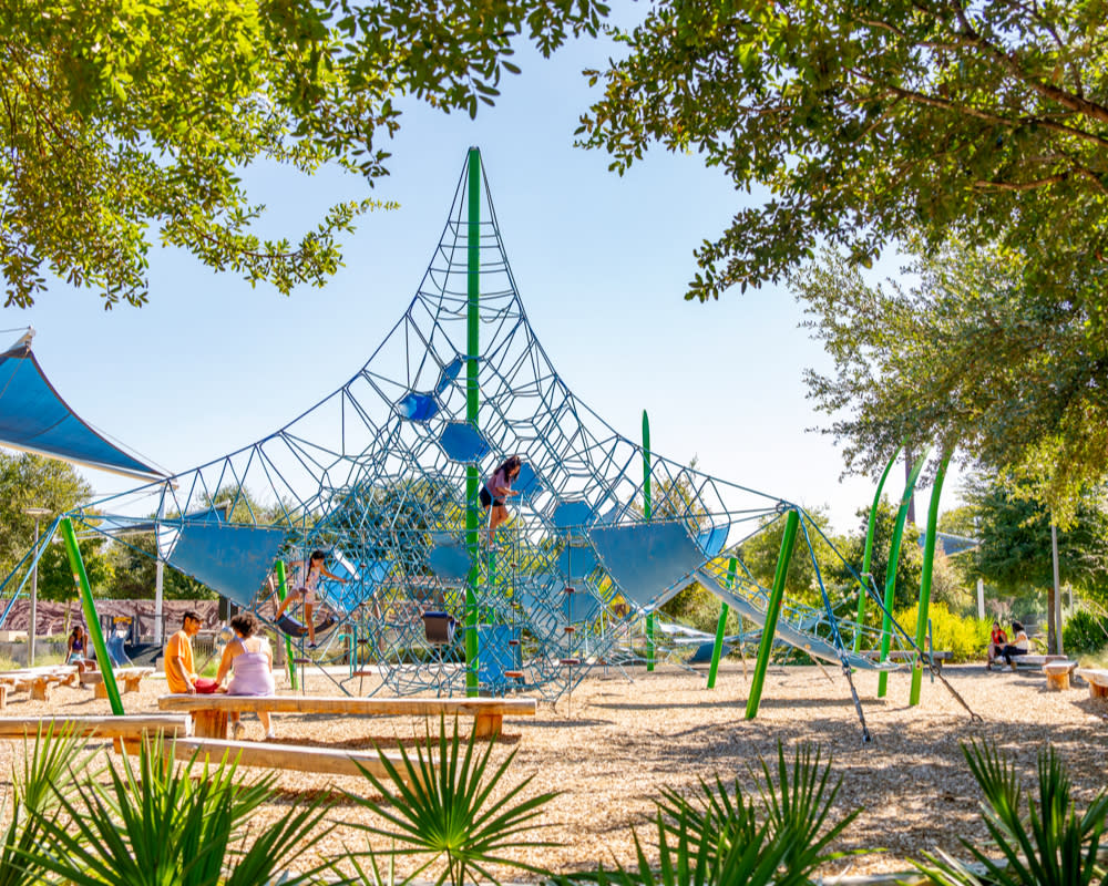 Two children climbing on a netted playscape while their parents watch.