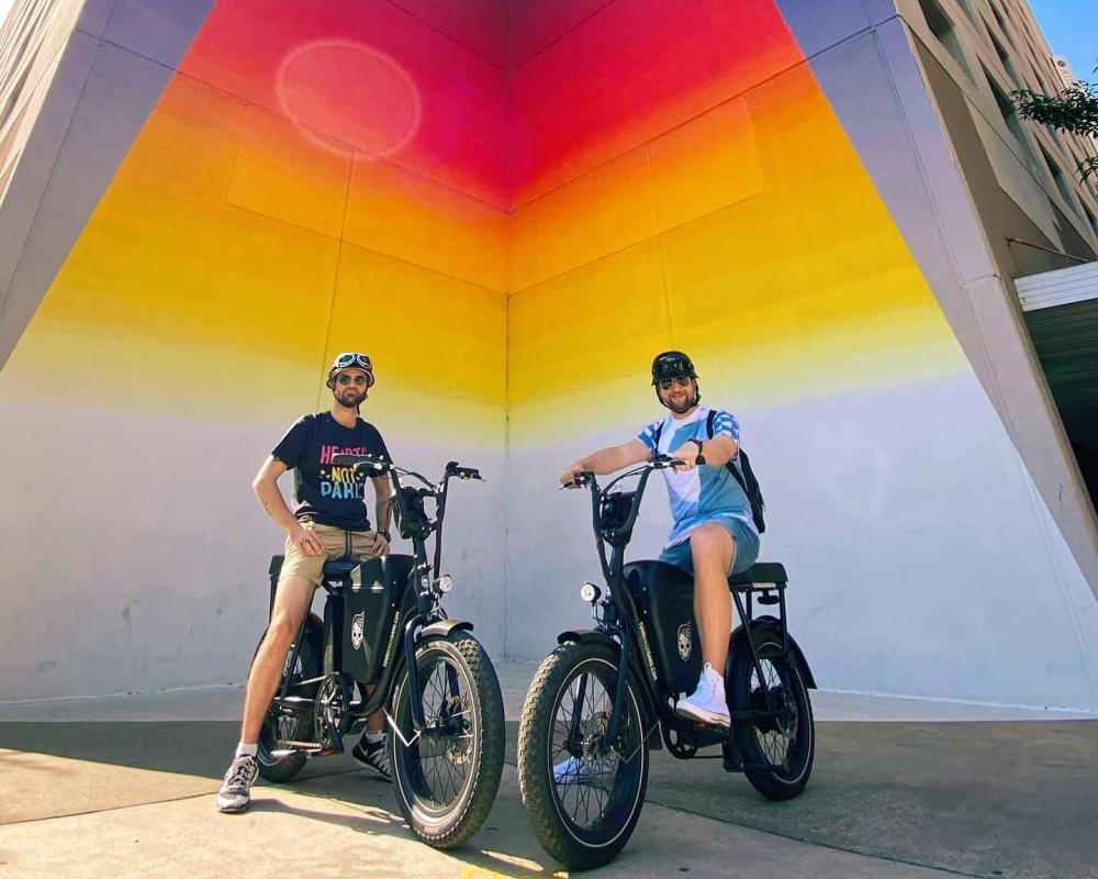Two men on Your Mini Biker Gang tour bikes in front of the Tau Ceti mural in downtown Austin.