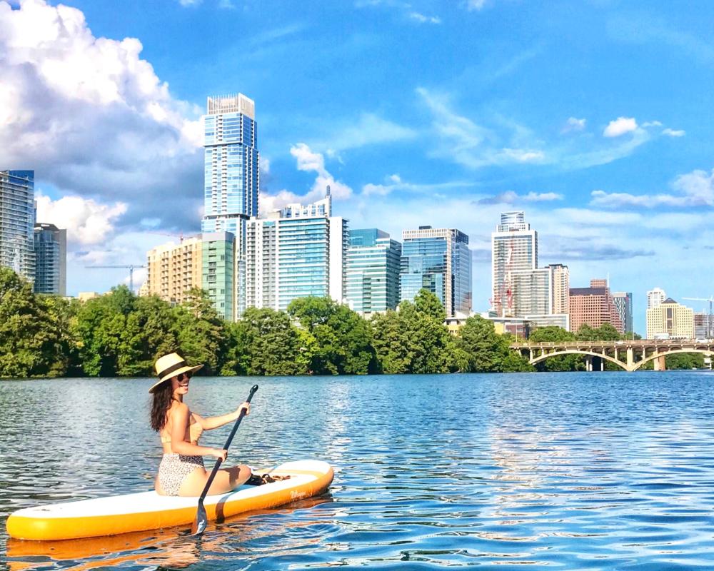 Girl in swimsuit and hat, sitting on paddle board on Lady Bird Lake looking towards the downtown skyline.