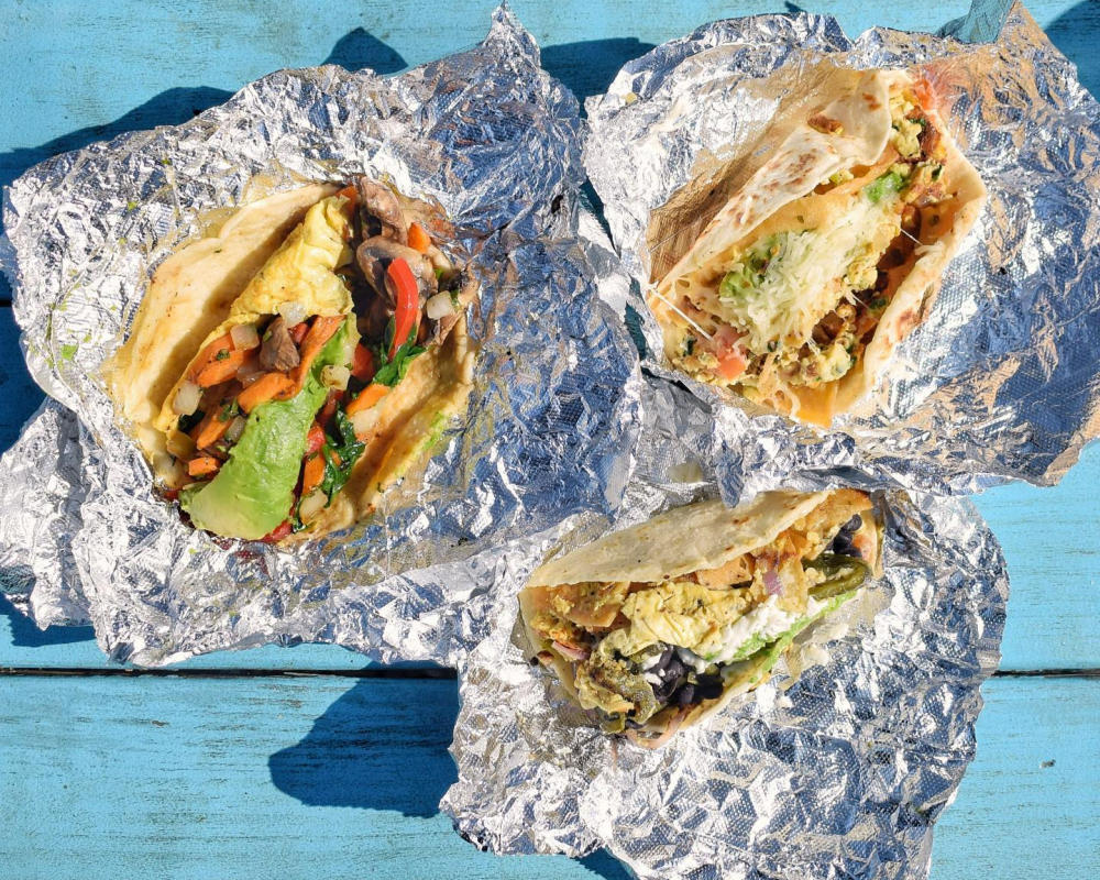 Three stuffed breakfast tacos in open foil on a blue picnic table top.
