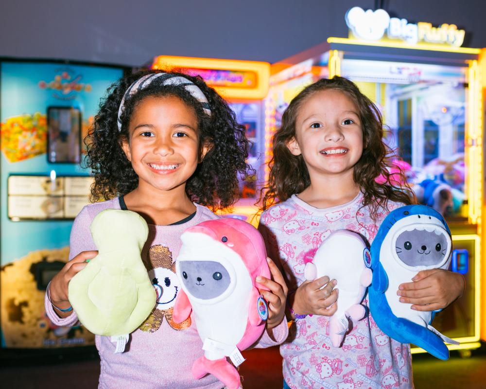 Two young girls standing in front of arcade machines, holding colorful plush animal toys.
