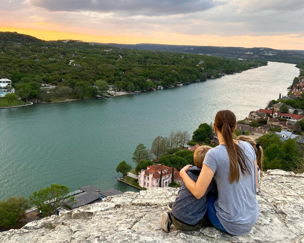 Image of a woman and child sitting atop Mount Bonnell facing away from the camera towards Lake Austin.