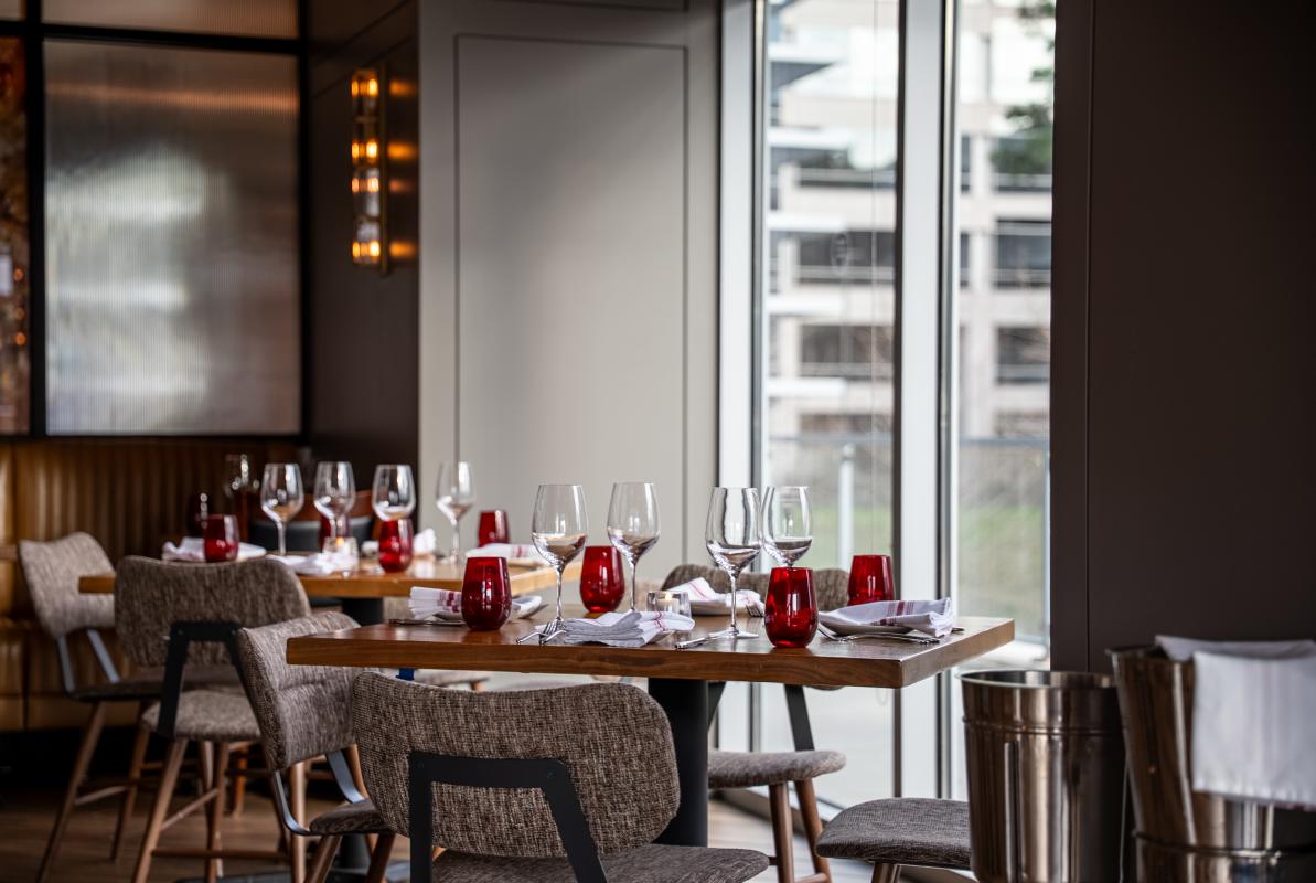 Two tables for four are seated beside a floor-to-ceiling window overlooking The Waterway. The tables are set with four wine glasses, water glasses, and red-and-white cloth napkins placed atop a simple white plate. The chairs are small but plush. The lighting is dim, but the room is lit with overcast daylight.