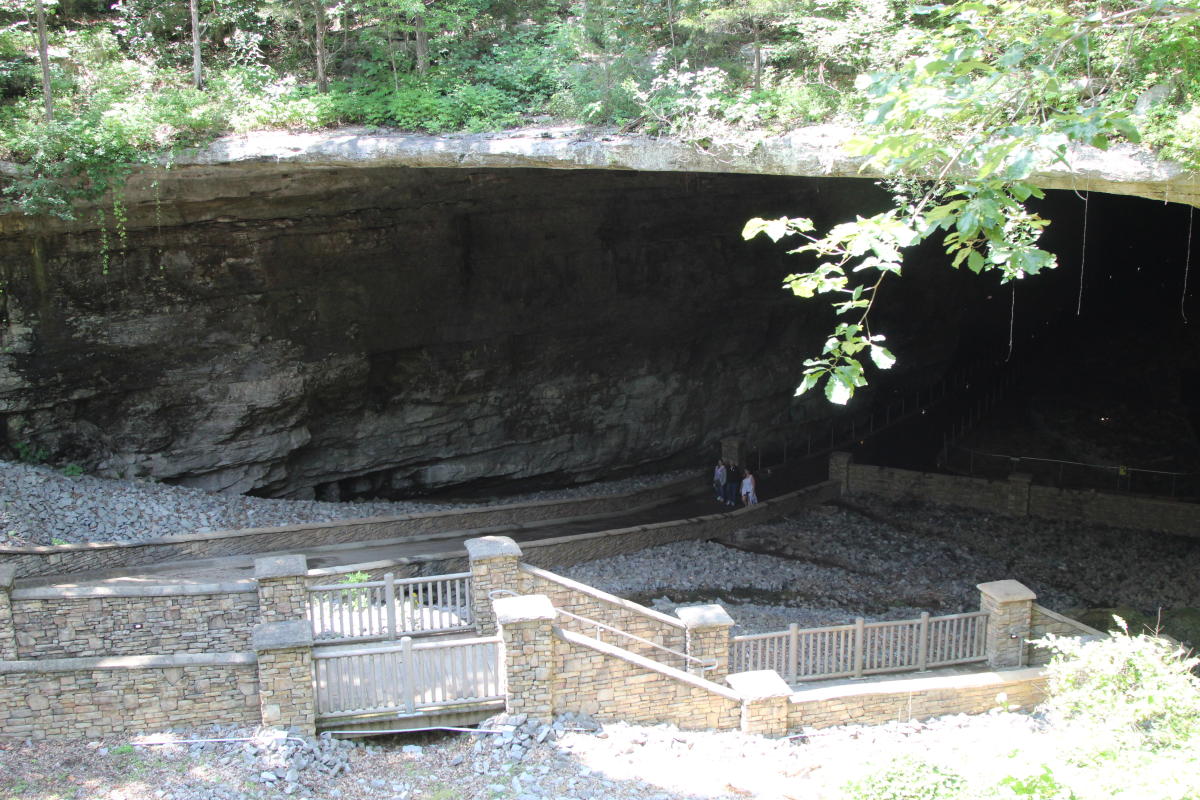 Very large opening to the mouth of a cave with stairs in the front and grass growing up above the opening.