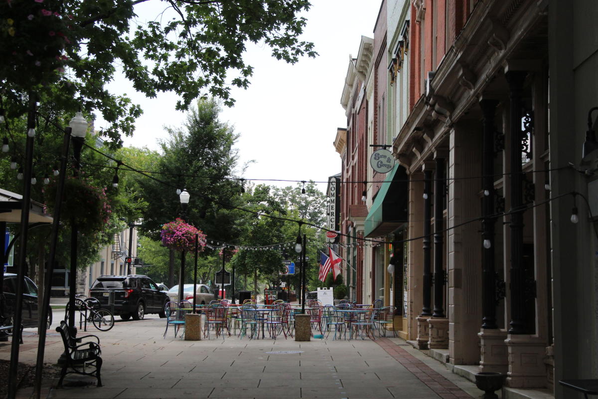 The photo shows a tree-lined downtown street with historic brick buildings on the right and hanging flower baskets on black lampposts. A row of colorful café tables and chairs sits in the middle of the wide sidewalk, strung with overhead lights that add a festive feel. Parked cars and green trees can be seen in the background, with storefront signs and flags adding detail to the street.