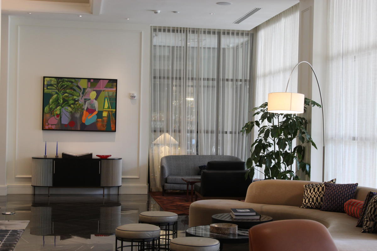 The photo shows a stylish hotel lobby with polished dark floors and soft natural light filtering through tall white curtains. A curved beige sofa with patterned pillows and round tables sits in the foreground, while a leafy green plant and tall floor lamp add warmth to the space. In the background, there are additional couches and a console table with candles and a colorful framed painting above it. The overall look is modern, clean, and inviting.