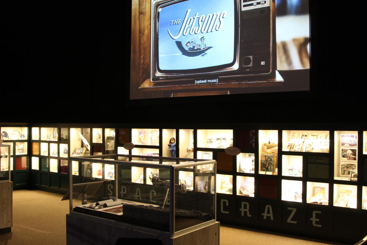 Dark room with lit displays of space memorabilia and a television screen playing the Jetsons cartoon at the top.
