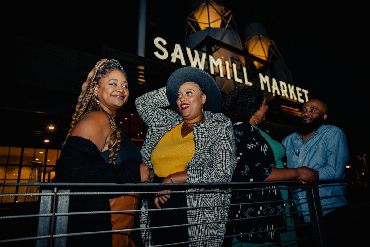 Four people laugh outside the Sawmill Market sign