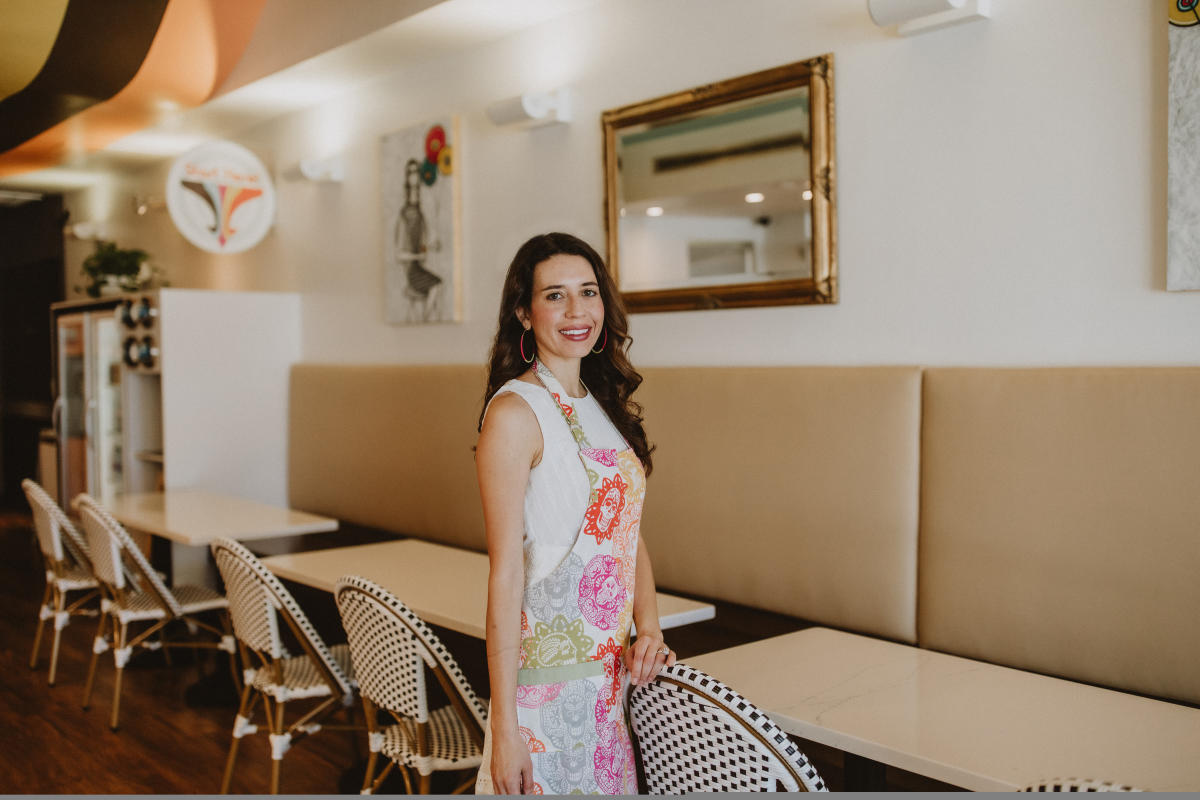 Camilla Dominguez, wearing a pink Dia de Los Muertos-themed apron, stands in center frame in her dessert shop, Olo Dessert Studio.