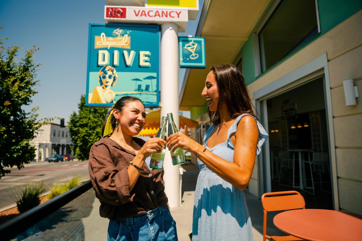 Two women "cheers" their bottles in front of a sign for the Imperial Dive.