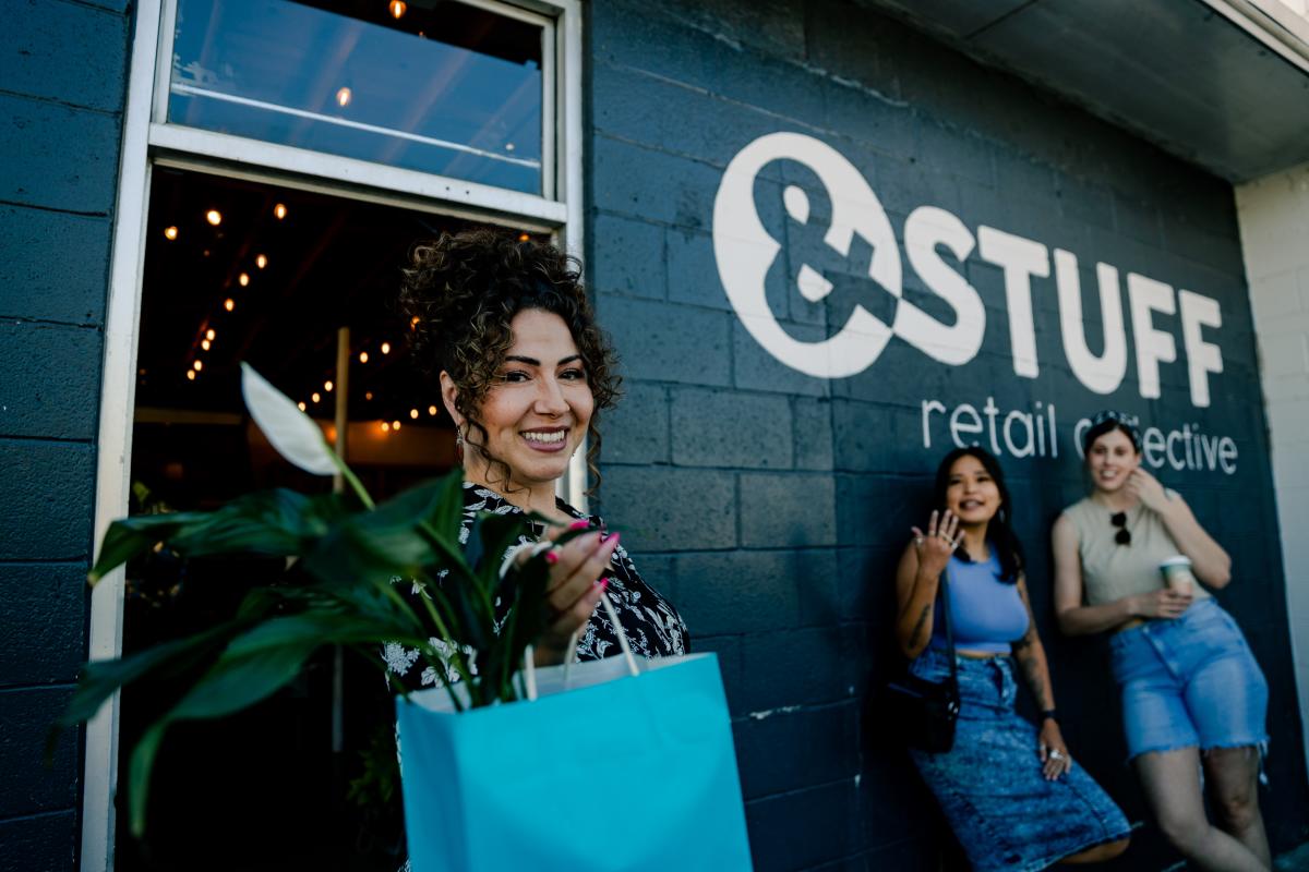 Two women watch as another woman holds up a bag of goods from And Stuff