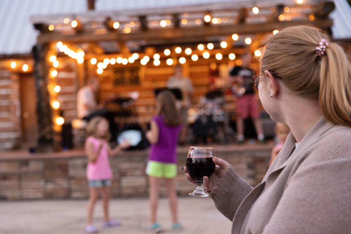 Two girls dance to music in front of a small stage while a mother looks on with a glass of beer in hand.