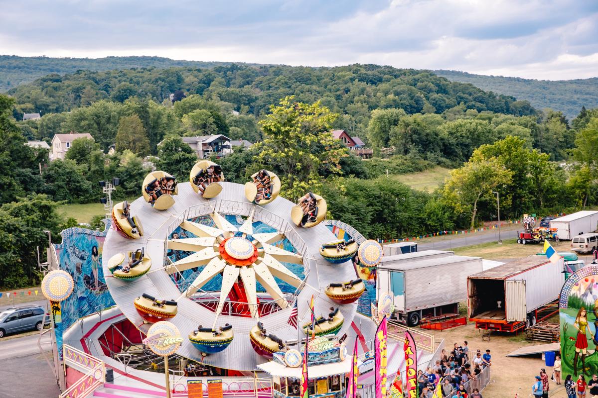 People riding a spinning carnival ride at the Allegany County Fair, with colorful flags, booths, and wooded hills in the background.