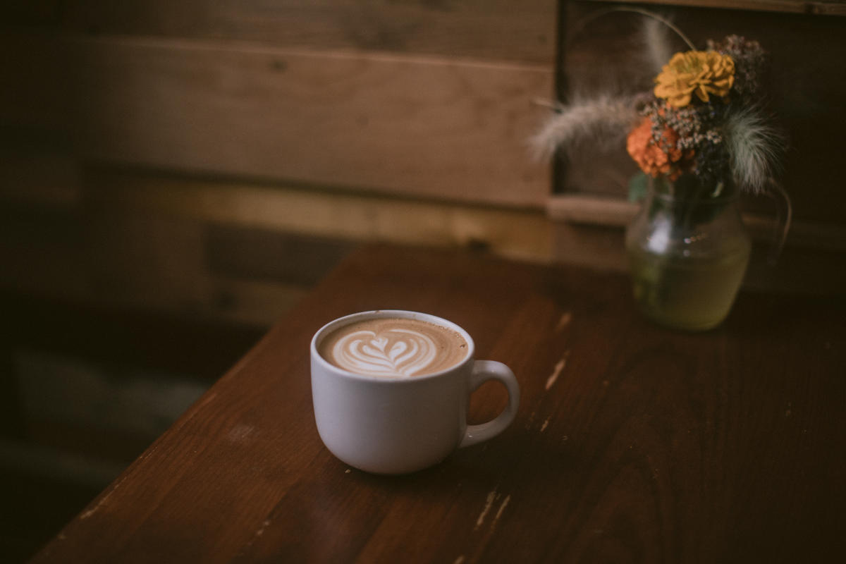 A latte served in a white cup sits on a table next to a vase with orange and yellow flowers.