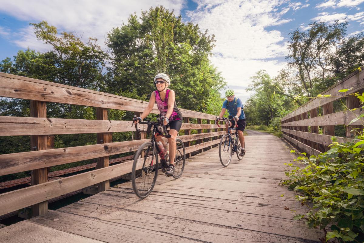 Two bikers cross a wooden bridge surrounded by green trees along the Great Allegheny Passage.