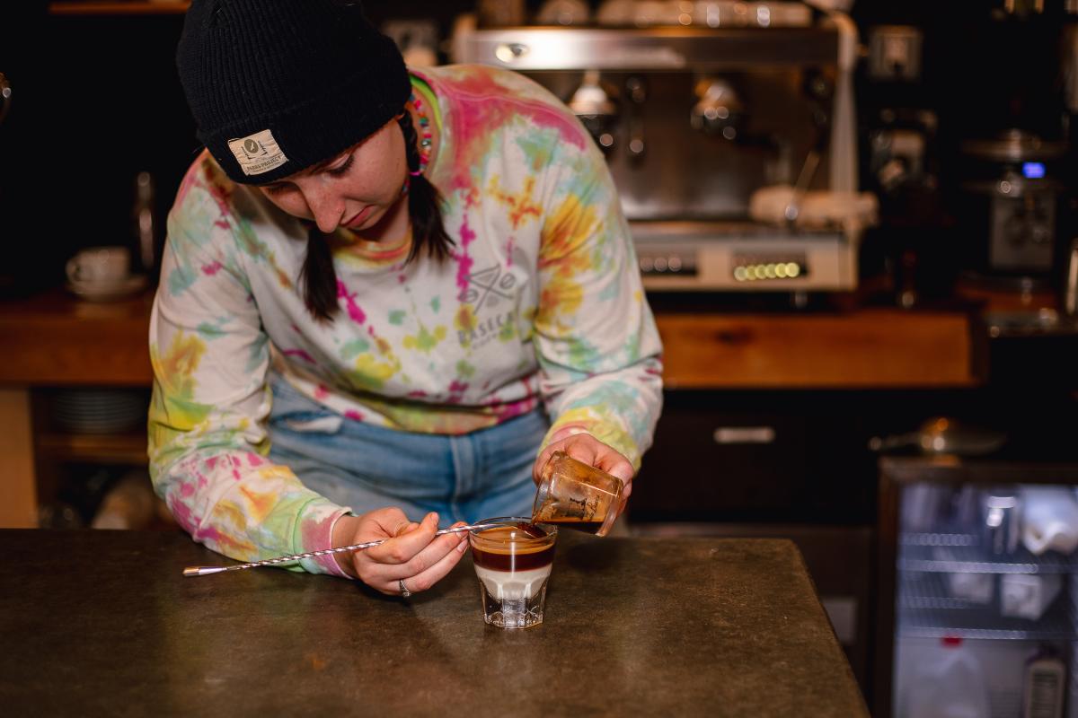 A barista in a tie-dye long sleeve and black beanie on her head, pours espresso into a glass.