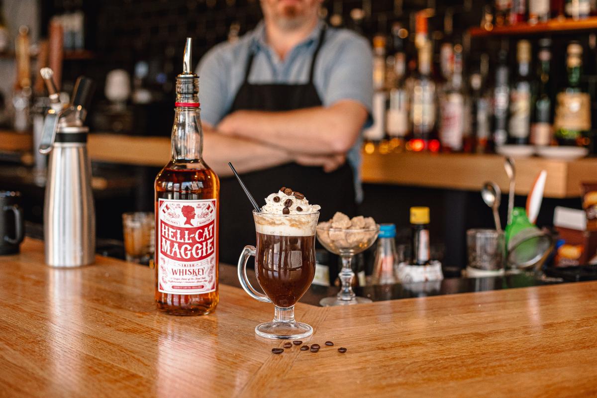 A coffee drink with whipped cream and coffee beans sits on a bar next to a bottle of Hell Cat liquor.