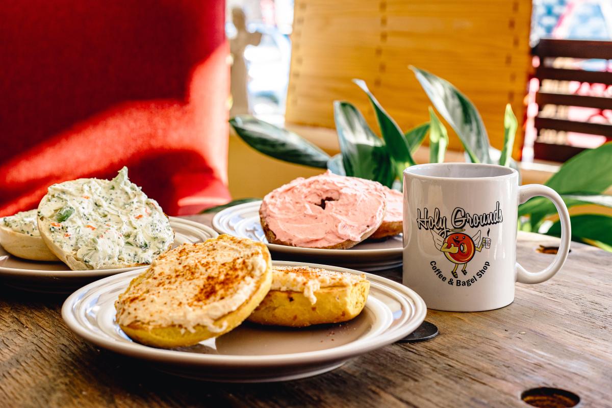 Three plates, each with a bagel and cream cheese, sit on a table next to a cup of coffee.