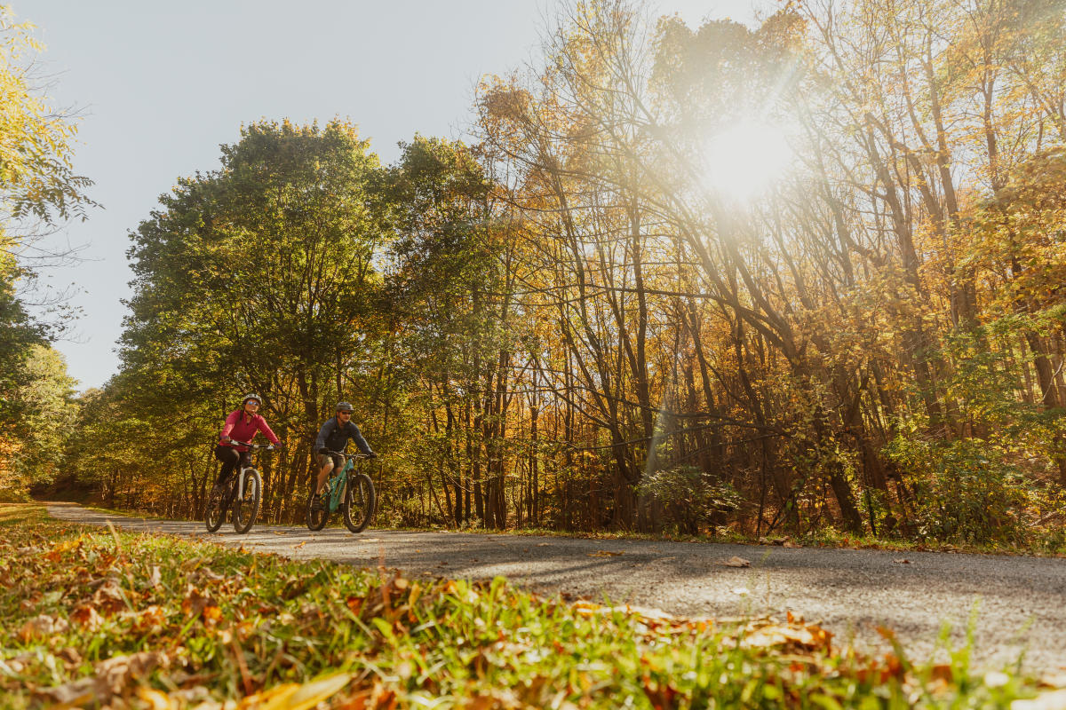 A man and a woman are each on their own bike. peddling along the Great Allegheny Passage in the fall.