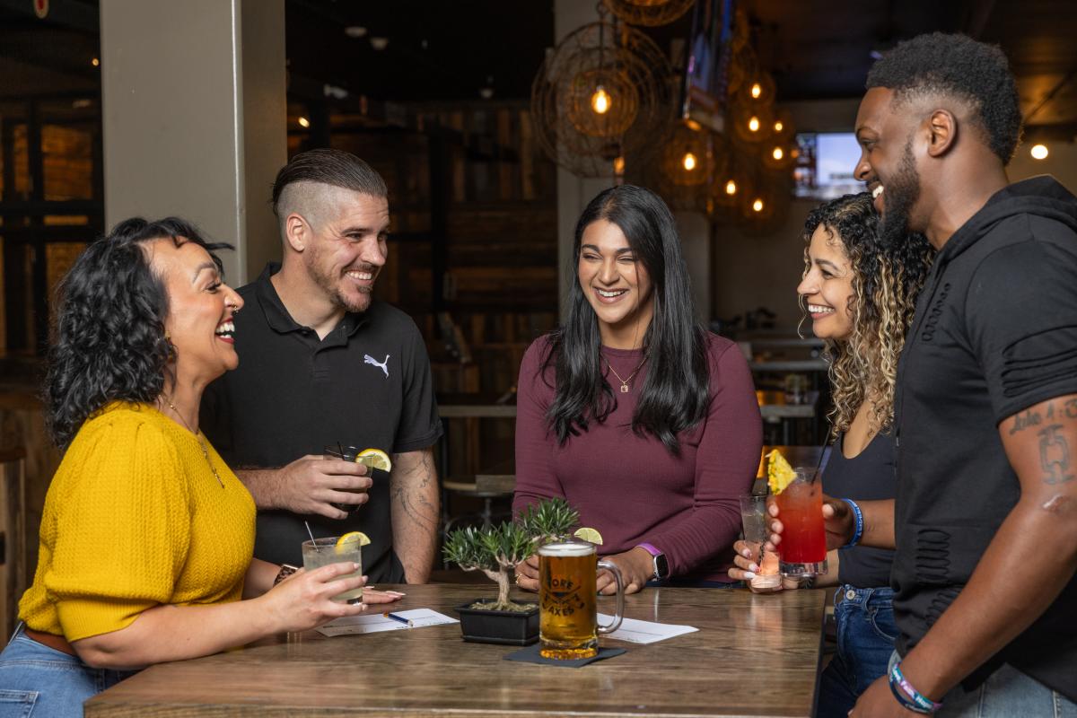 A group of adults stand around a high top bar table drinking cocktails.
