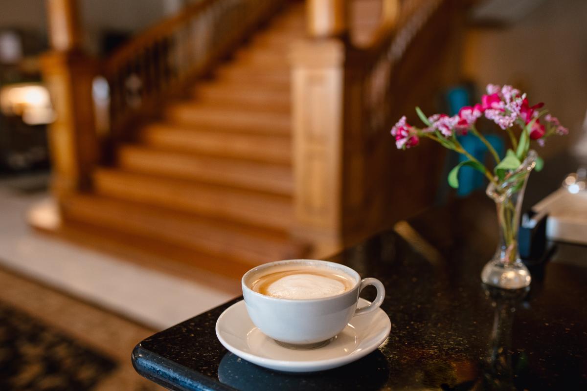 A latte in white cup sits on the edge of a table next to a vase with purple flowers.