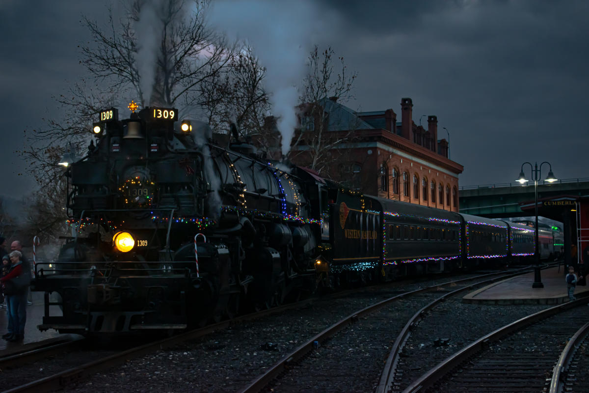 A steam engine decorated with Christmas lights and a holiday wreath on the engine is parked in front of a historic brick depot at night.