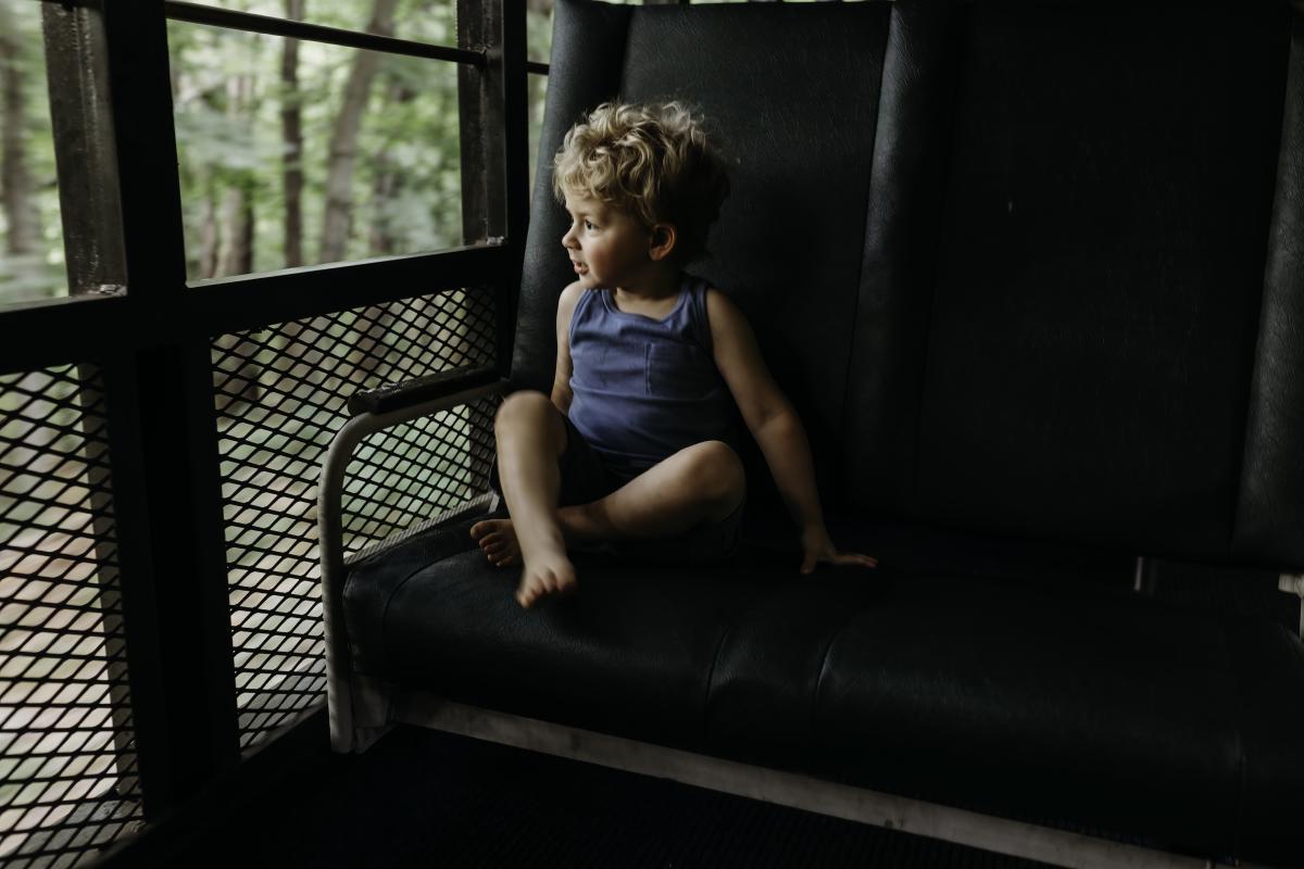 A blonde haired boy in a blue shirt and shorts stares out the window of a train.