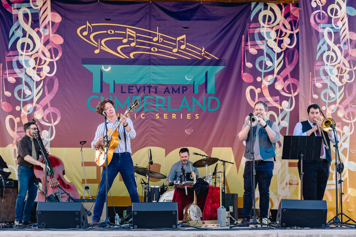 Full band on stage playing music in front of a colorful backdrop that says "Levitt AMP Cumberland Music Series."