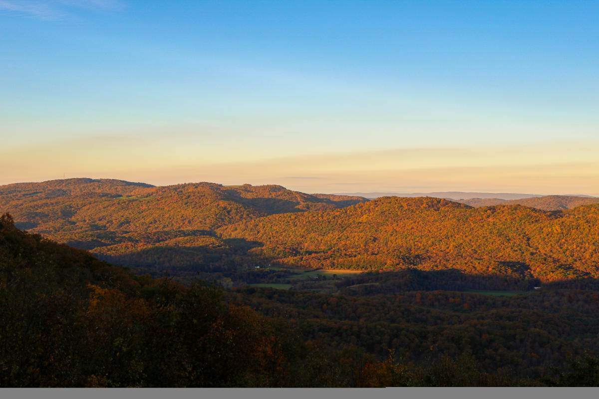 A mountain range is bright with shades of fall color, and the sky is a shade of blue as the sunrises behind the mountains