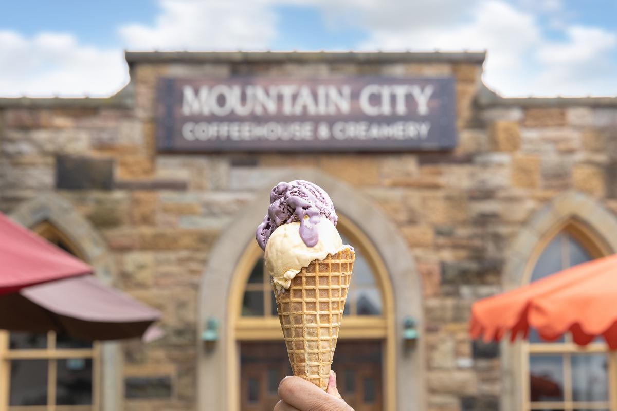 A hand holds up a waffle cone with blueberry and vanilla ice cream in front of a stone mason building with a sign that says Mountain City Coffee House and Creamery.