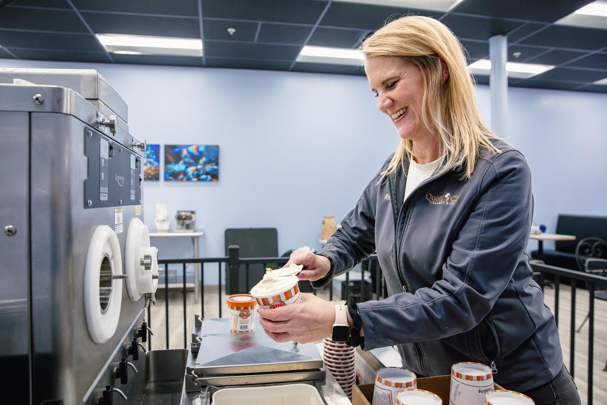 A blonde haired woman scopes ice cream from a manufacturing size ice cream machine into pint cups.