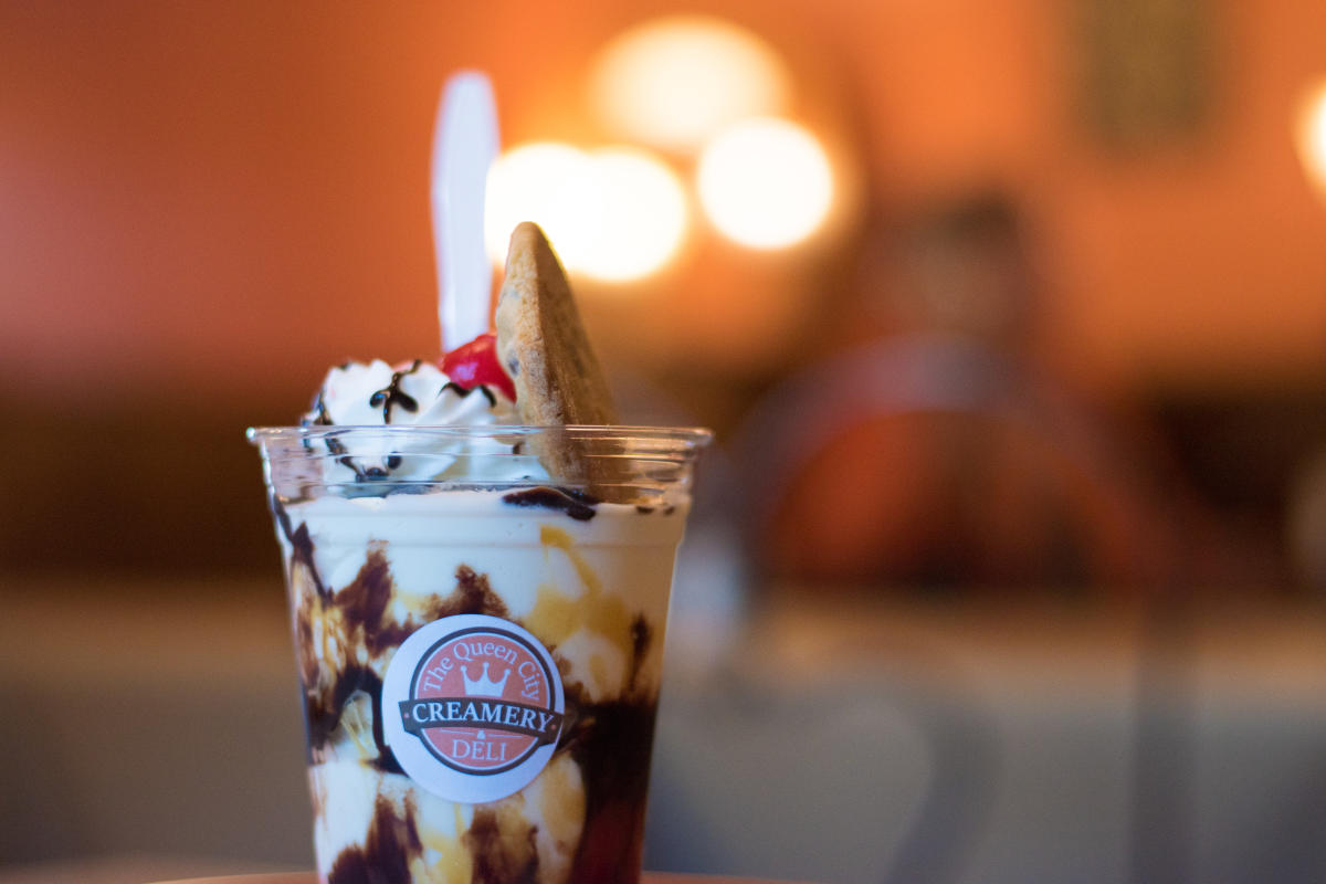 A cup of Queen City Creamery vanilla ice cream with hot fudge and topped with whip cream, a cheery, and a large chocolate chip cookie.