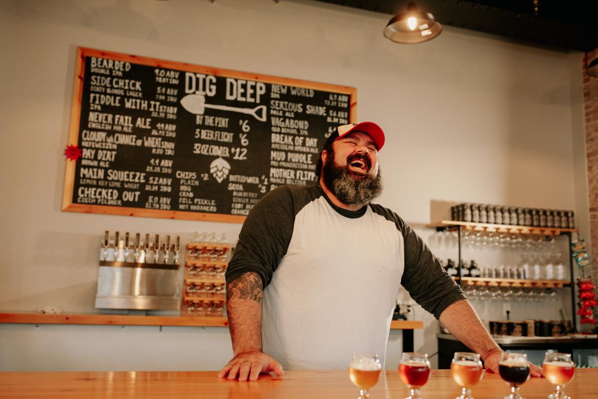 A man in a white shirt with green sleeves leans on a bar laughing over a flight of beers.