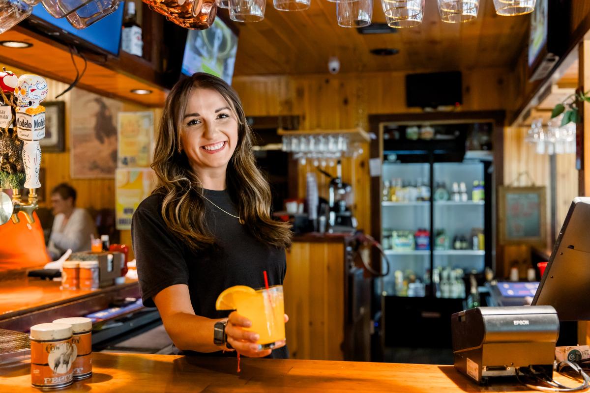 A female bartender hands over an orange crush trail cocktail at the bar.
