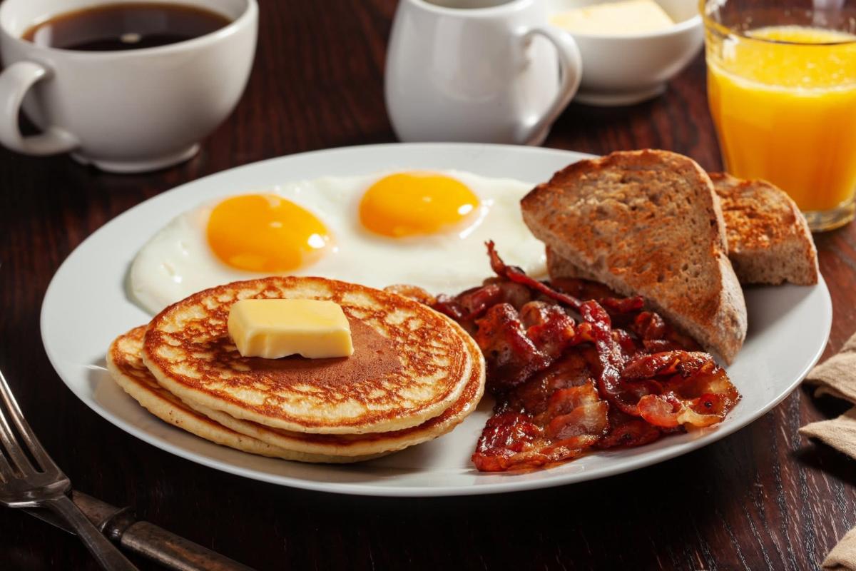 Plate of breakfast food with pancakes topped with butter, two sunny-side-up eggs, bacon, toast, coffee, and orange juice.