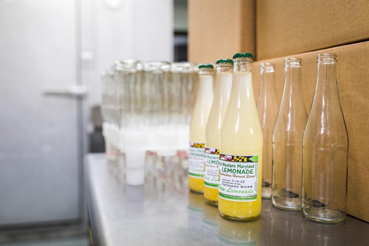 Empty glass bottles are lined up on a counter with the front three filled with lemoande and a label, all ready to be packaged in nearby cardboard boxes.