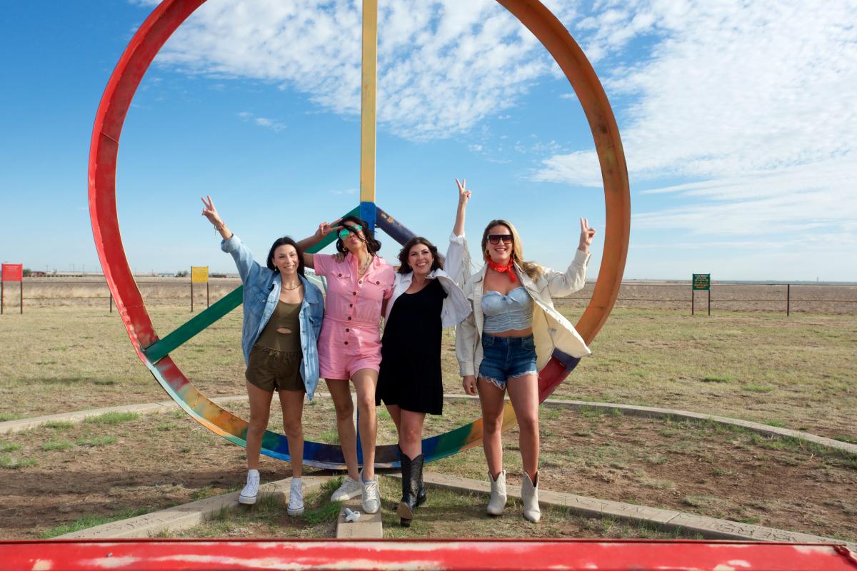 Four women pose in front of a large, colorful peace sign sculpture under a bright West Texas sky. Dressed in playful, casual outfits with touches of Western flair—like denim jackets, boots, and a red bandana—they flash peace signs and big smiles. The wide-open plains stretch behind them, capturing the laid-back, free-spirited vibe of Amarillo’s quirky roadside art scene.