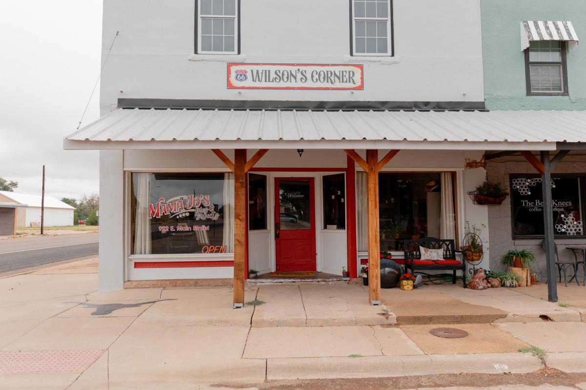A storefront along Route 66 featuring Wilson’s Corner, a small-town building with a red door and wooden pillars. To the left, Mama Jo’s Pies & More displays a neon “OPEN” sign in the window, while The Bee’s Knees coffee and boutique is visible on the right with flower pots and a cozy bench outside. The sidewalk and street hint at a quiet, welcoming Main Street atmosphere.