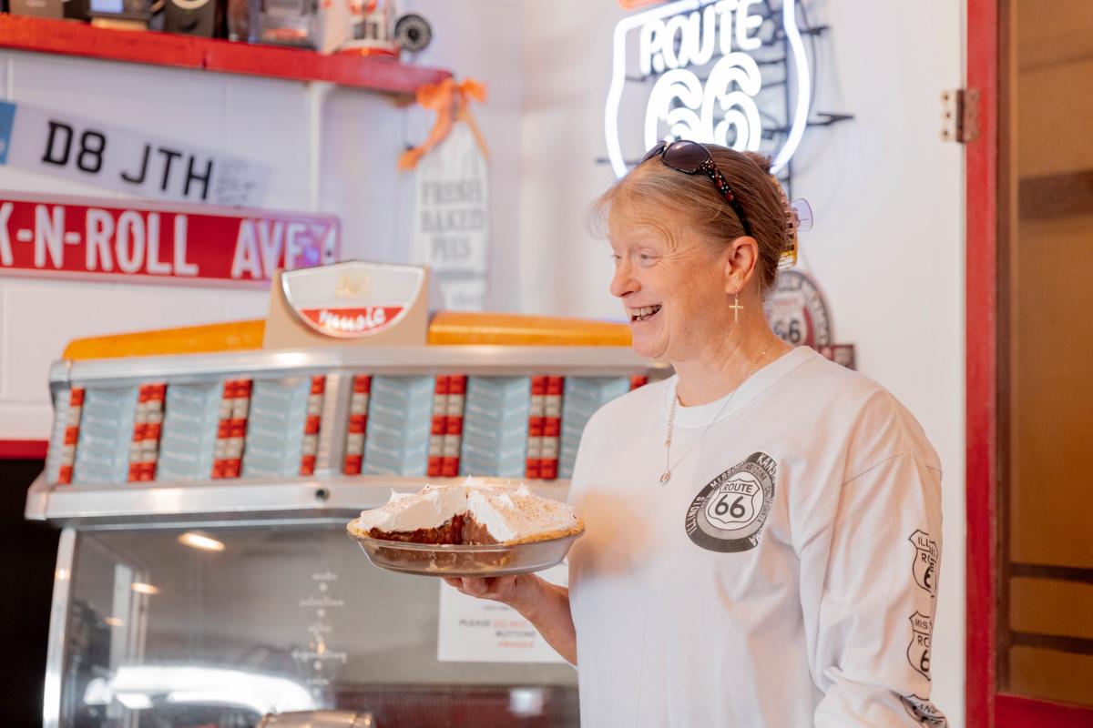 A smiling woman in a Route 66-themed diner holds a slice of pie topped with fluffy meringue. She wears a long-sleeve Route 66 shirt and stands in front of a vintage jukebox, with neon signs and retro memorabilia surrounding her, capturing the charm and nostalgia of Texas' iconic Mother Road stops.