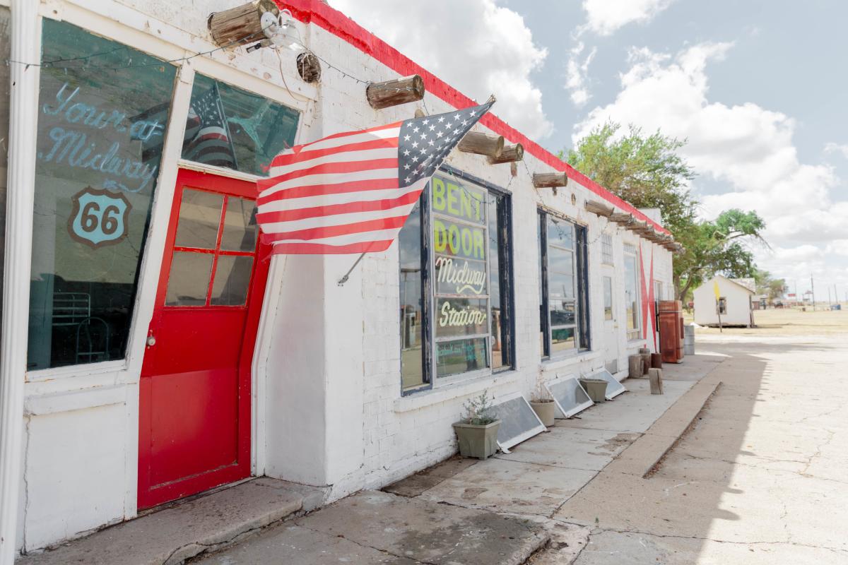 A weathered American flag flutters outside the Bent Door Midway Station, a white brick building with a bold red trim and a distinct slanted doorway. Set along historic Route 66, the structure’s vintage neon sign and retro charm evoke the spirit of classic road trips through West Texas.