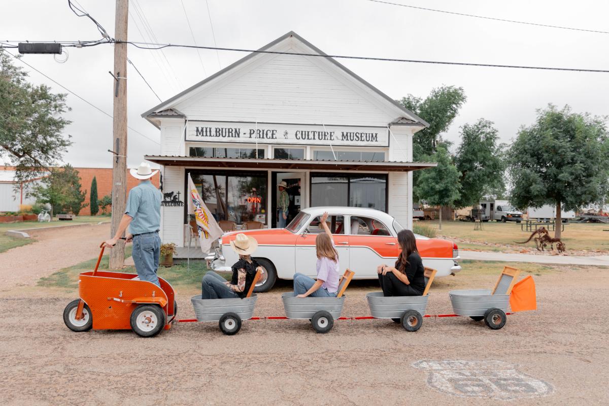 A man in a cowboy hat pulls a small barrel train filled with smiling riders past the Milburn-Price & Culture Museum, a white clapboard building along Route 66. A vintage orange-and-white car is parked out front, adding retro flair.