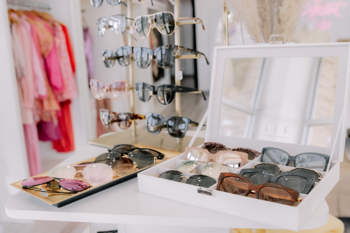 Photo of sunglasses in a case and clothing racks in the background with pink and white blouses