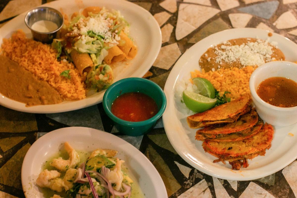 Plat of mexican food, plate one features agua chile, plate two is flautas covered in cheese served with rice and beans, plate three is tacos de birria served with rice and beans and consume at El Manatial